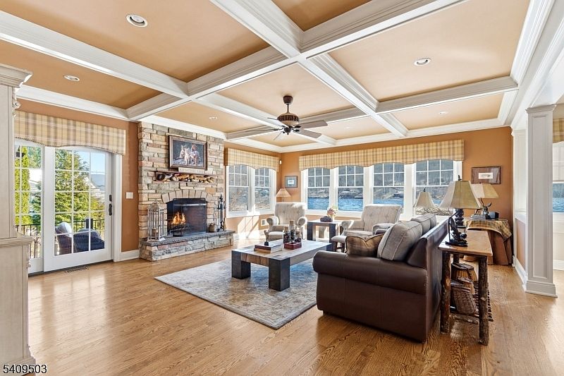 Coffered great room with hardwood floors, a stone fireplace under a custom mantle, white framed lake-view windows, and a French door opening onto the pavilion